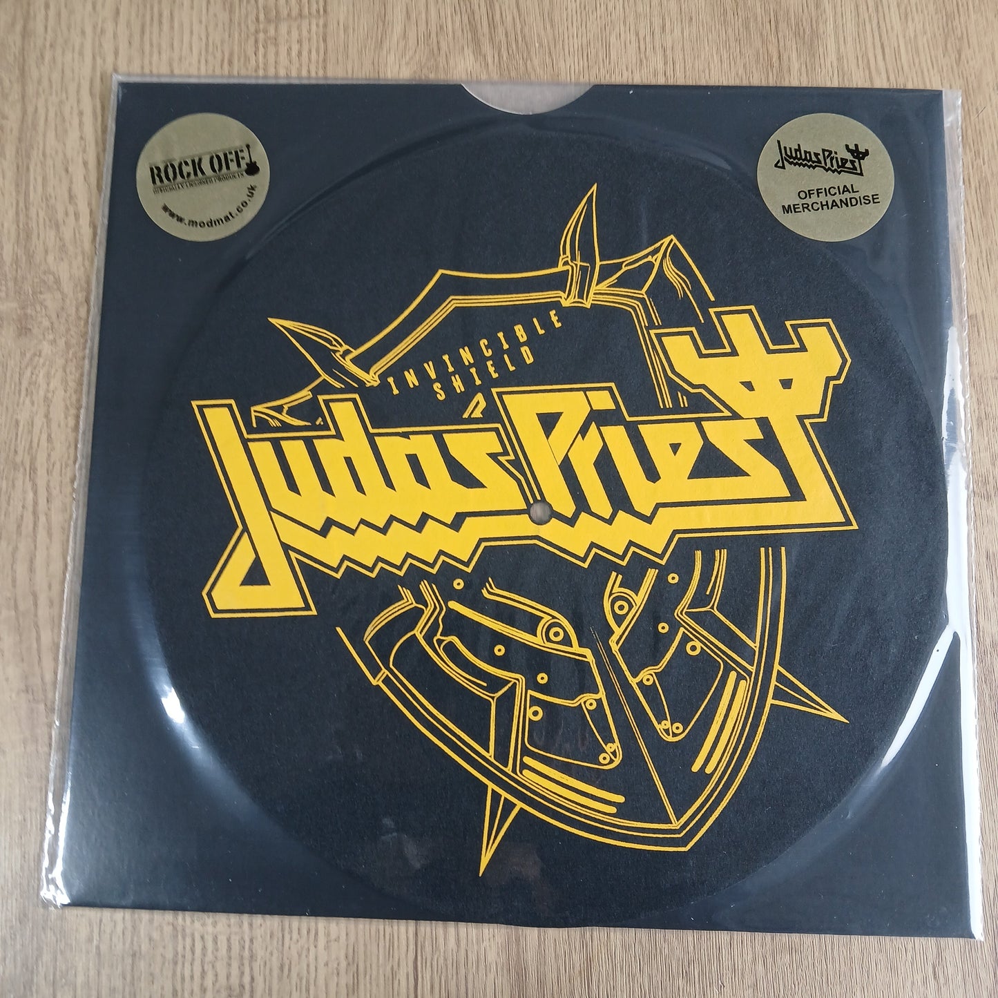 judas priest invincible shield  with yellow graphic design on a black background, placed on a wooden surface.