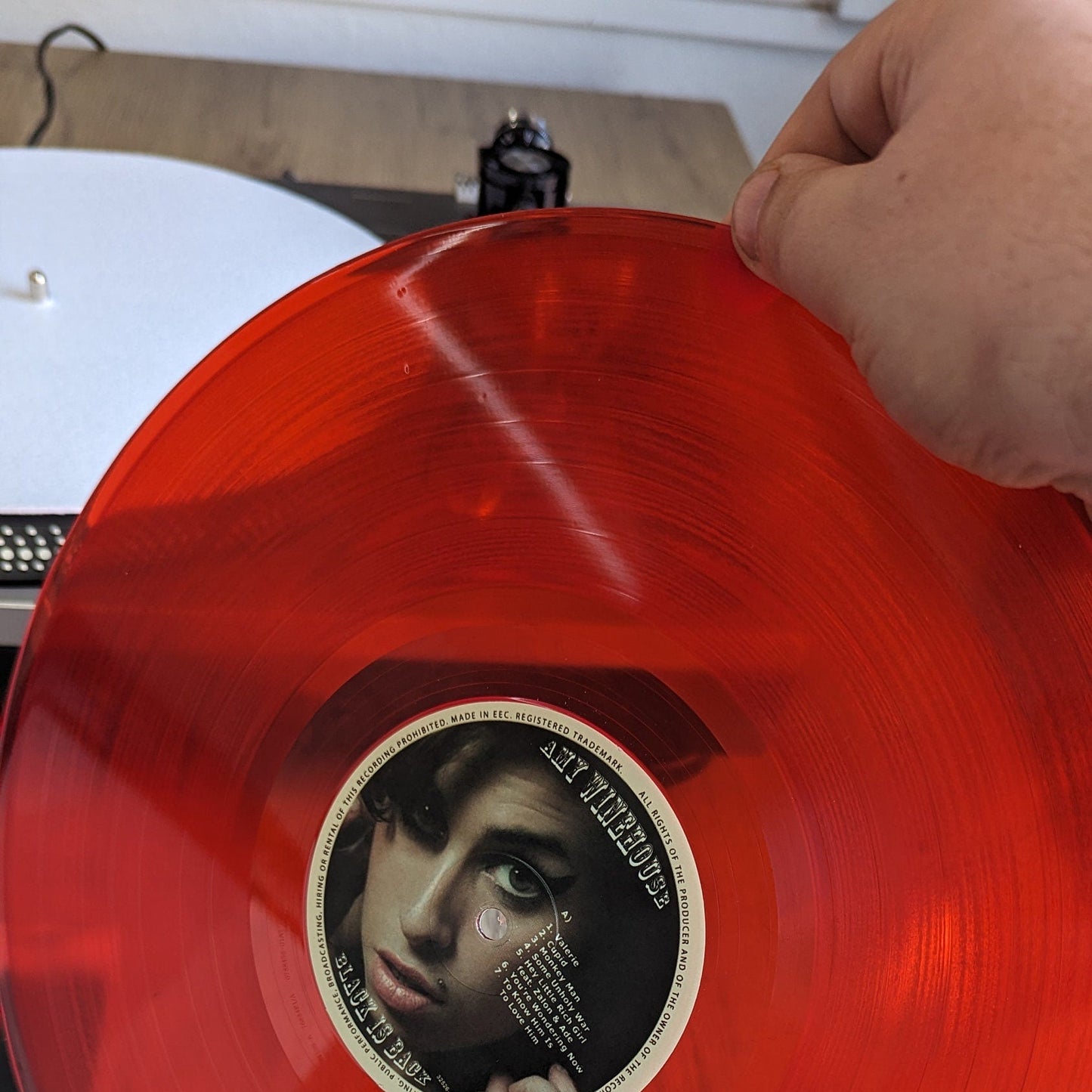 Red vinyl record with a label featuring a face of Amy winehouse, held by a hand on a wooden surface.