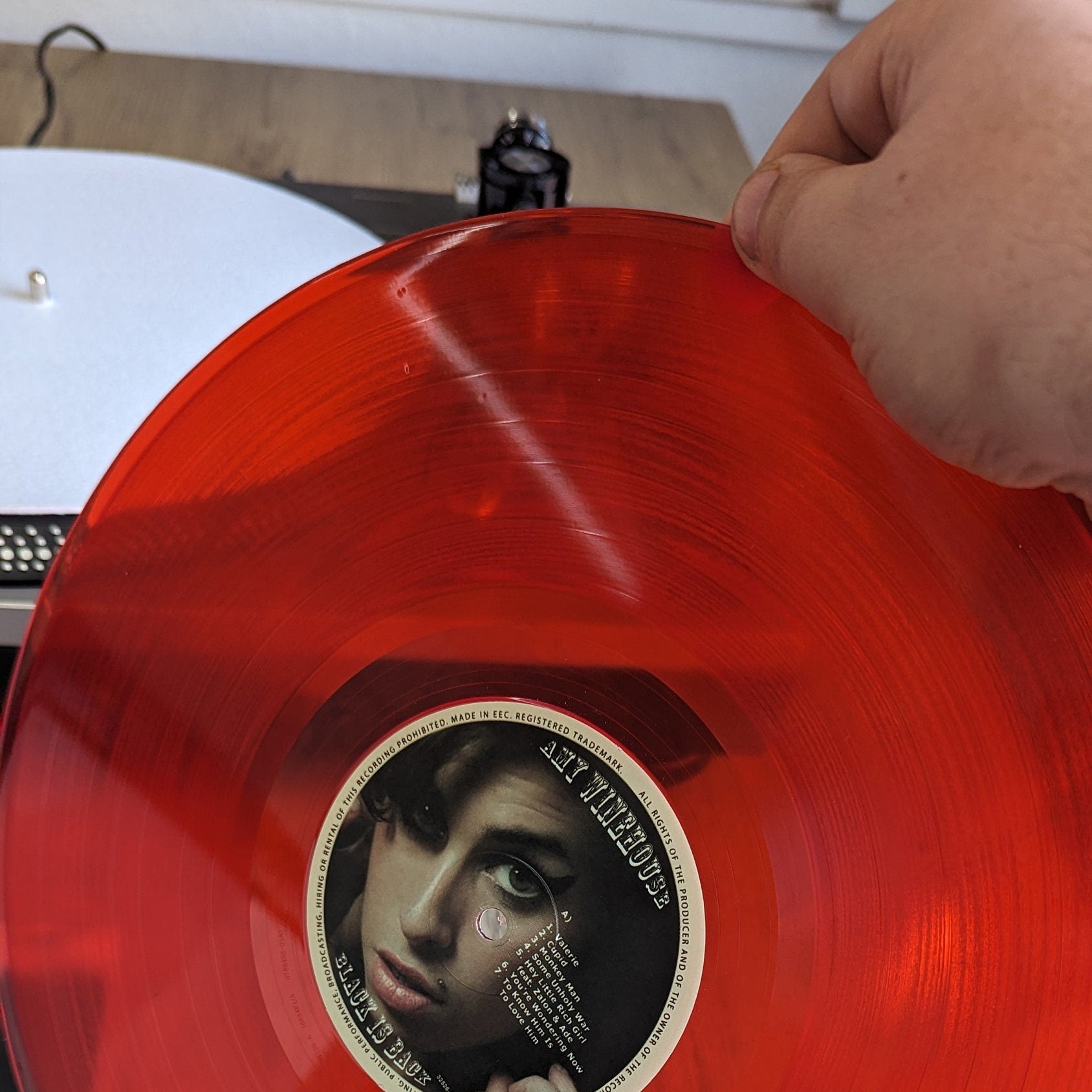 Red vinyl record with a label featuring a face of Amy winehouse, held by a hand on a wooden surface.
